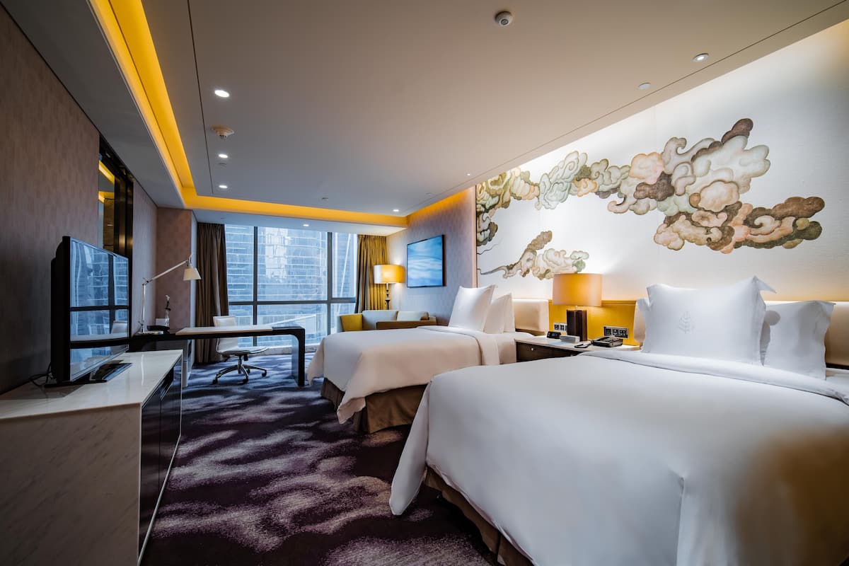A hotel room with two beds, a desk, a chair, a TV, and a large window with a city view.