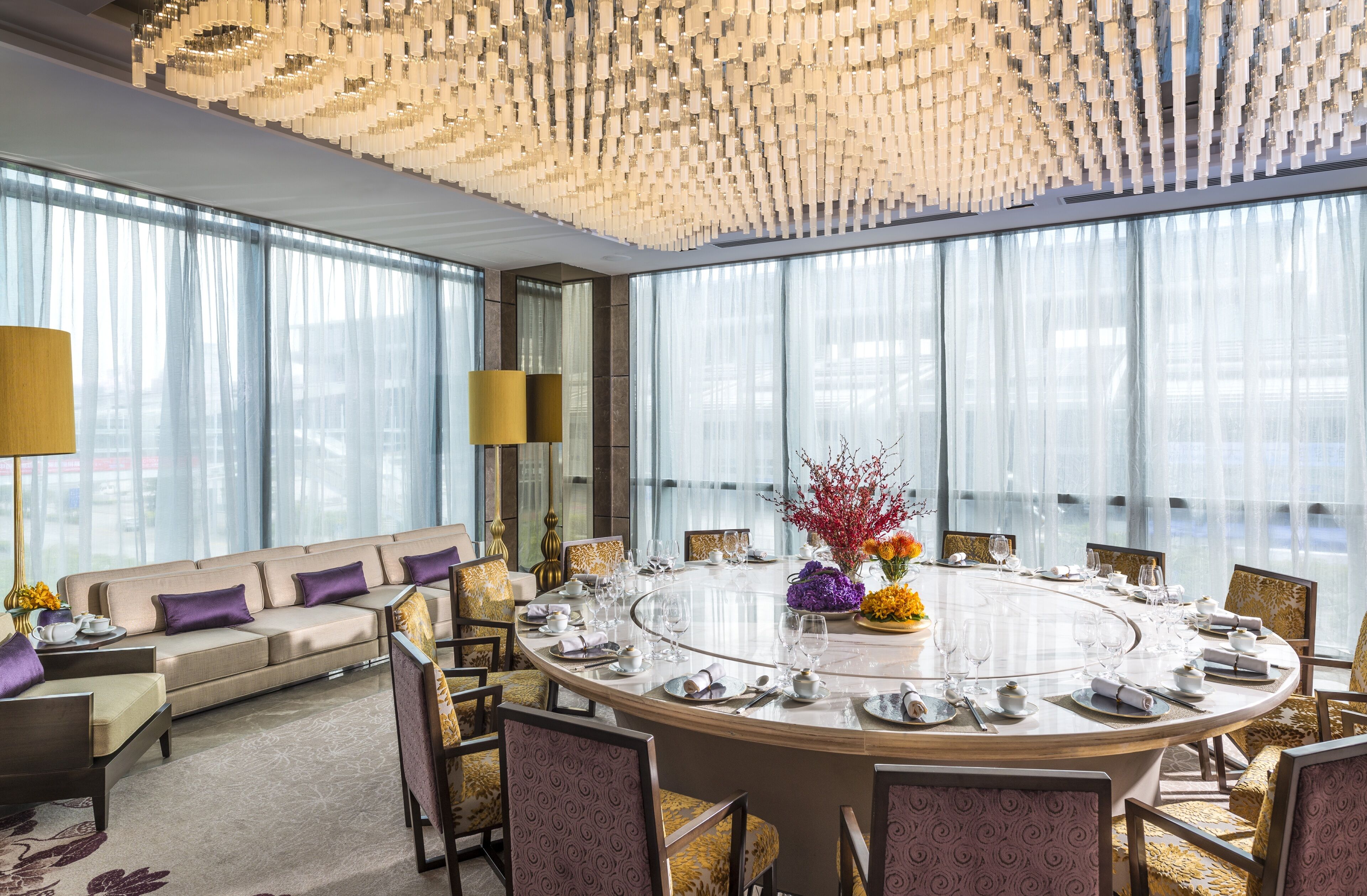 A modern hotel room with a round dining table set for six, a sofa, and a large window with sheer curtains.