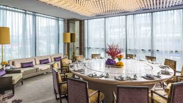 A modern hotel room with a round dining table set for six, a sofa, and a large window with sheer curtains.