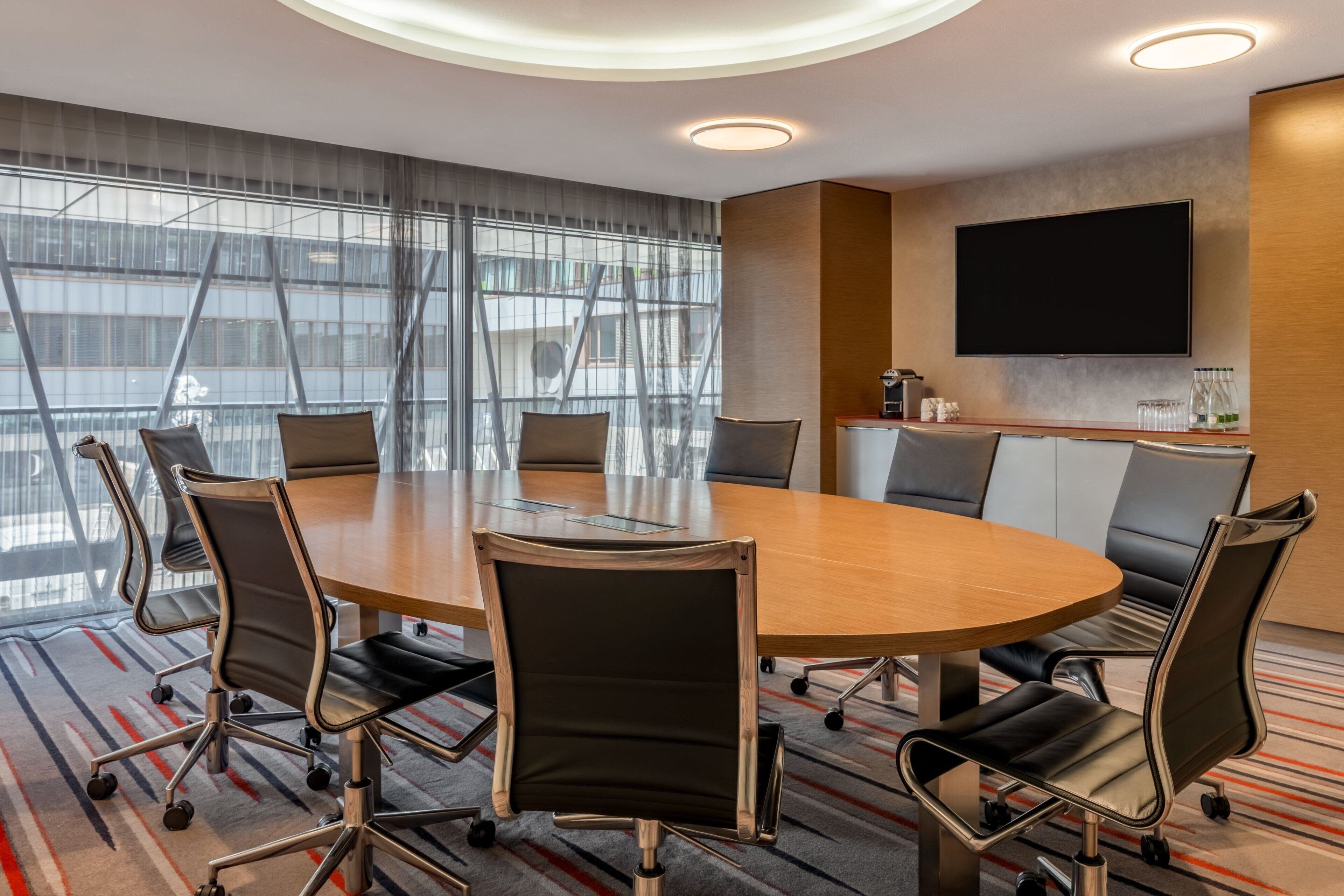 A conference room with a large oval table, black leather chairs, a flat-screen TV, and a large window with blinds.