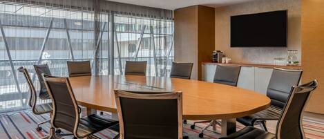 A conference room with a large oval table, black leather chairs, a flat-screen TV, and a large window with blinds.