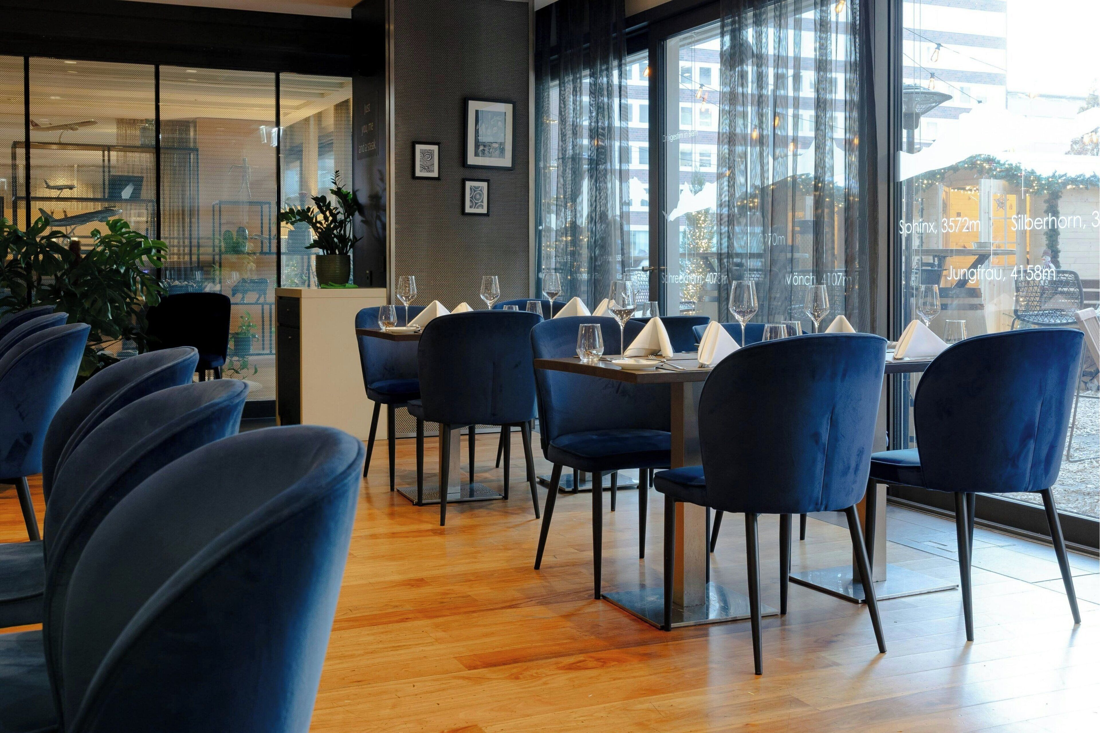 Un restaurante con sillas de terciopelo azul, mesas de madera y amplios ventanales con vistas a la ciudad.