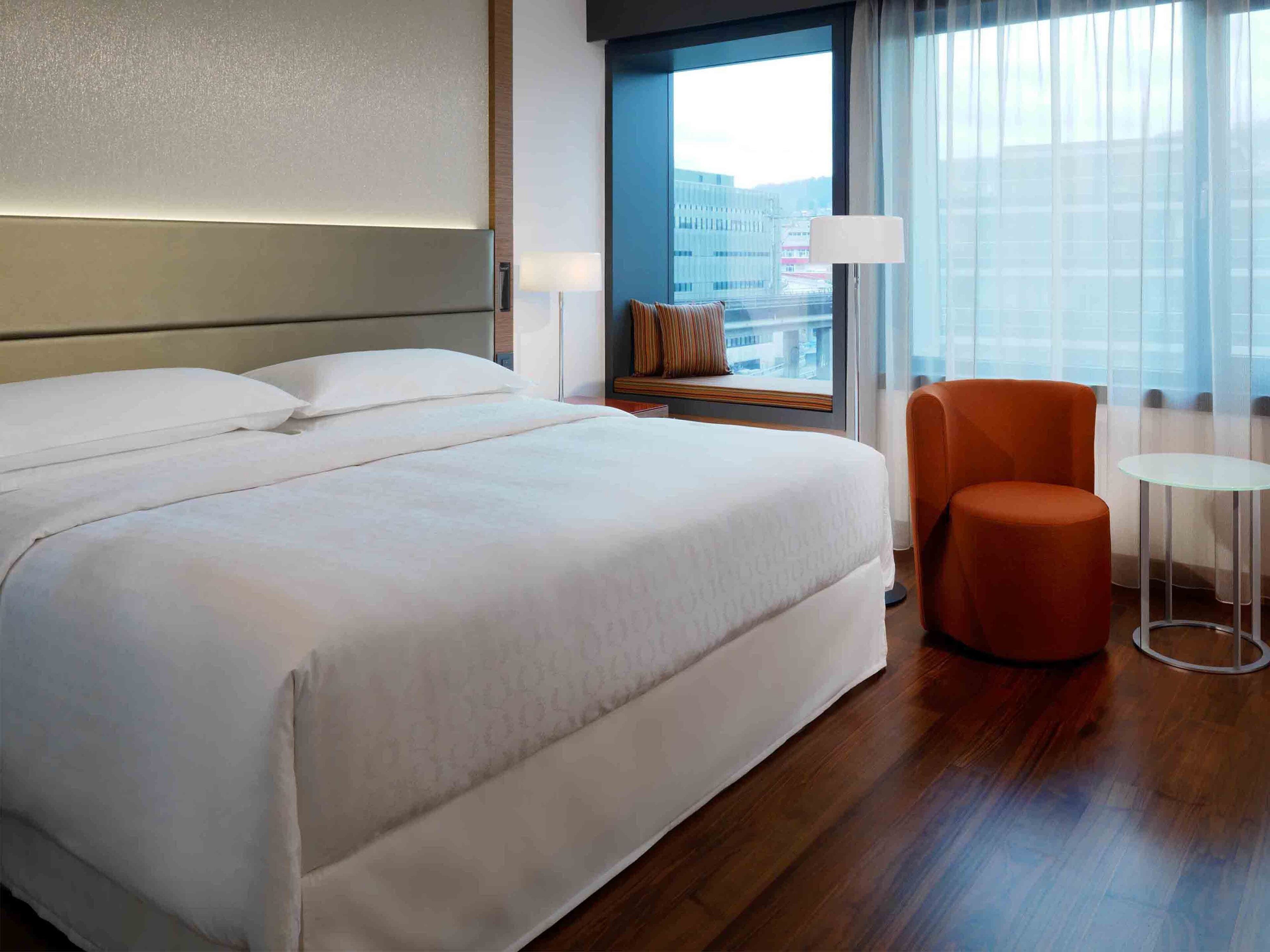 A hotel room with a large bed, a small table, an orange chair, a window with a view of buildings, and a lamp on a nightstand.