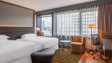 Ein Hotelzimmer mit zwei Betten, einem Schreibtisch, einem Stuhl, einem kleinen Tisch und Blick auf die Stadt.