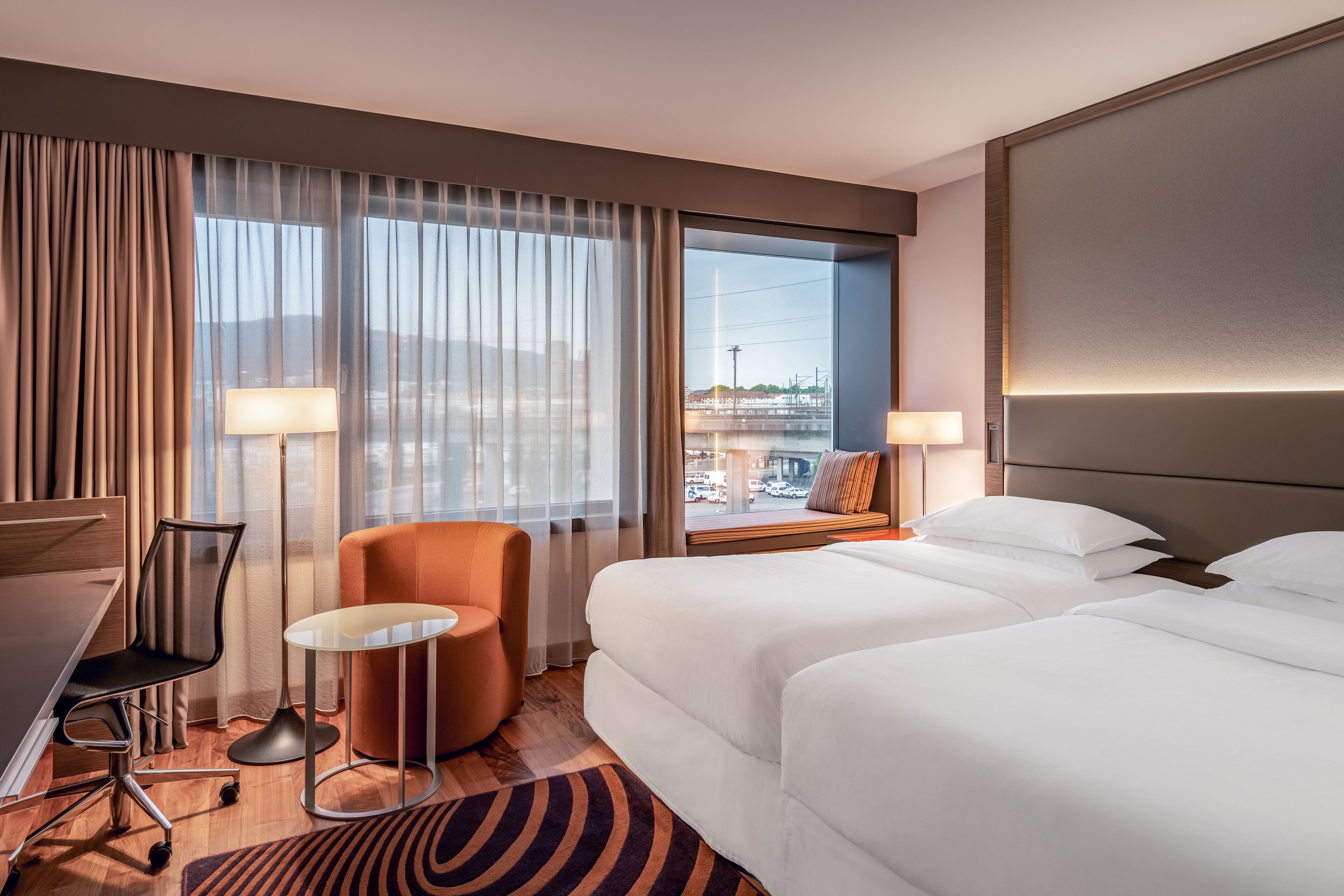 A hotel room with two beds, a desk, a chair, a small table, and a view of a cityscape through large windows.