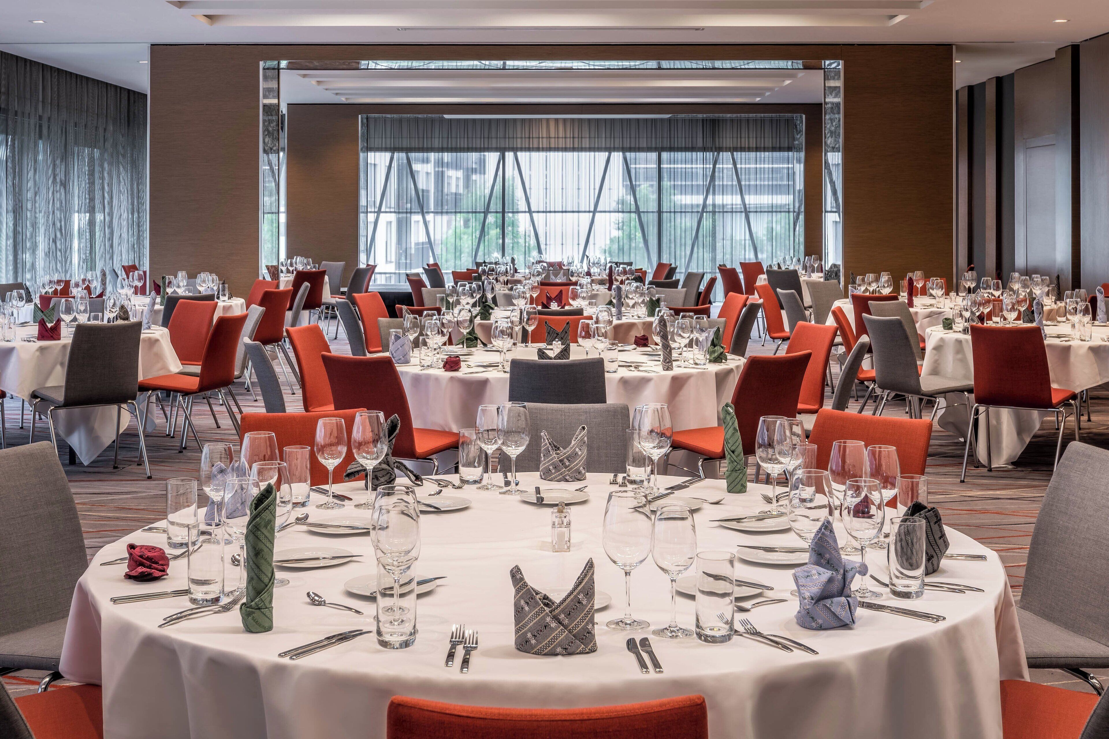 Un salón de banquetes con mesas redondas dispuestas para comer, con manteles blancos, sillas rojas y cubertería ordenada.