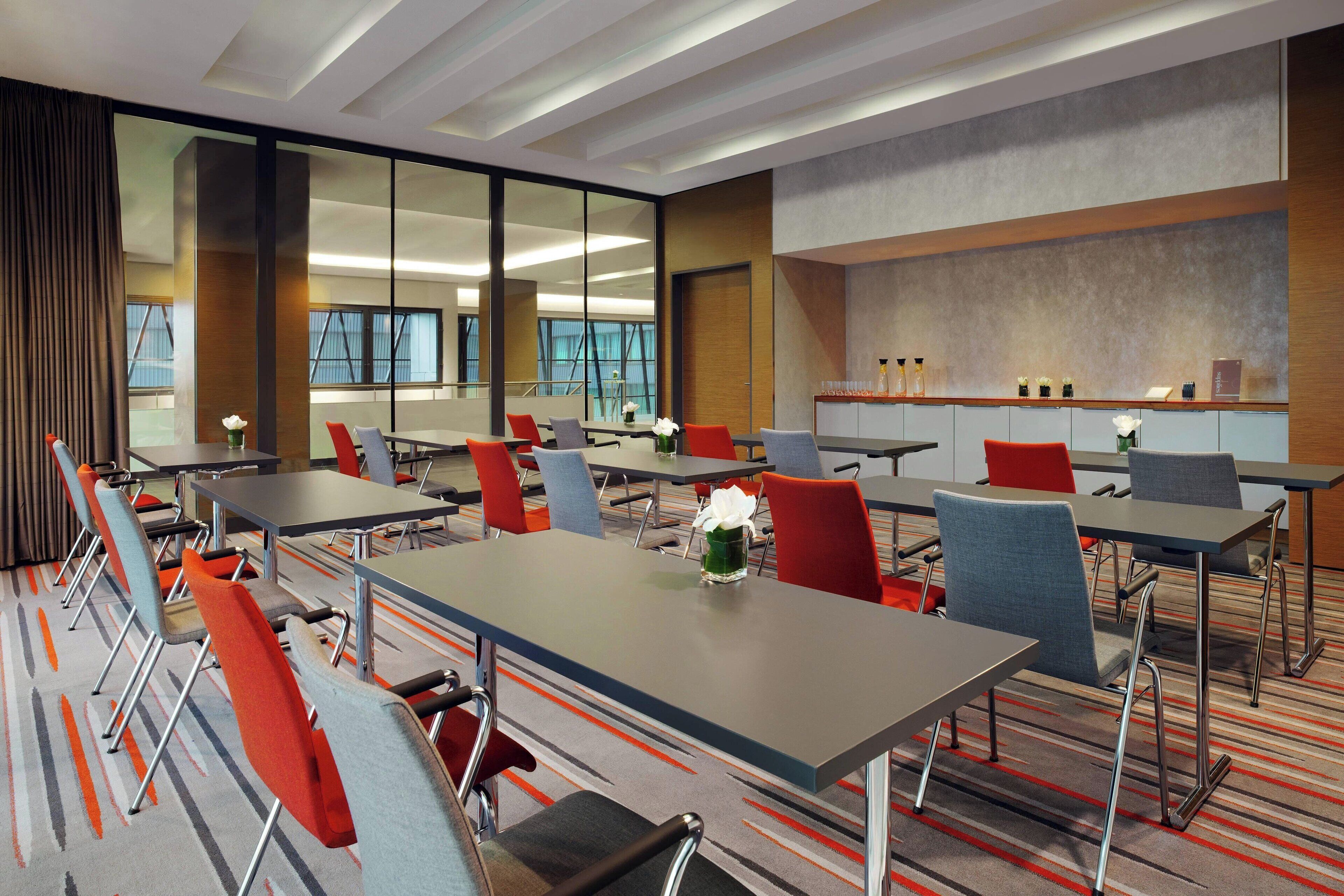A modern conference room with multiple tables and chairs, large windows, and a neutral color palette.