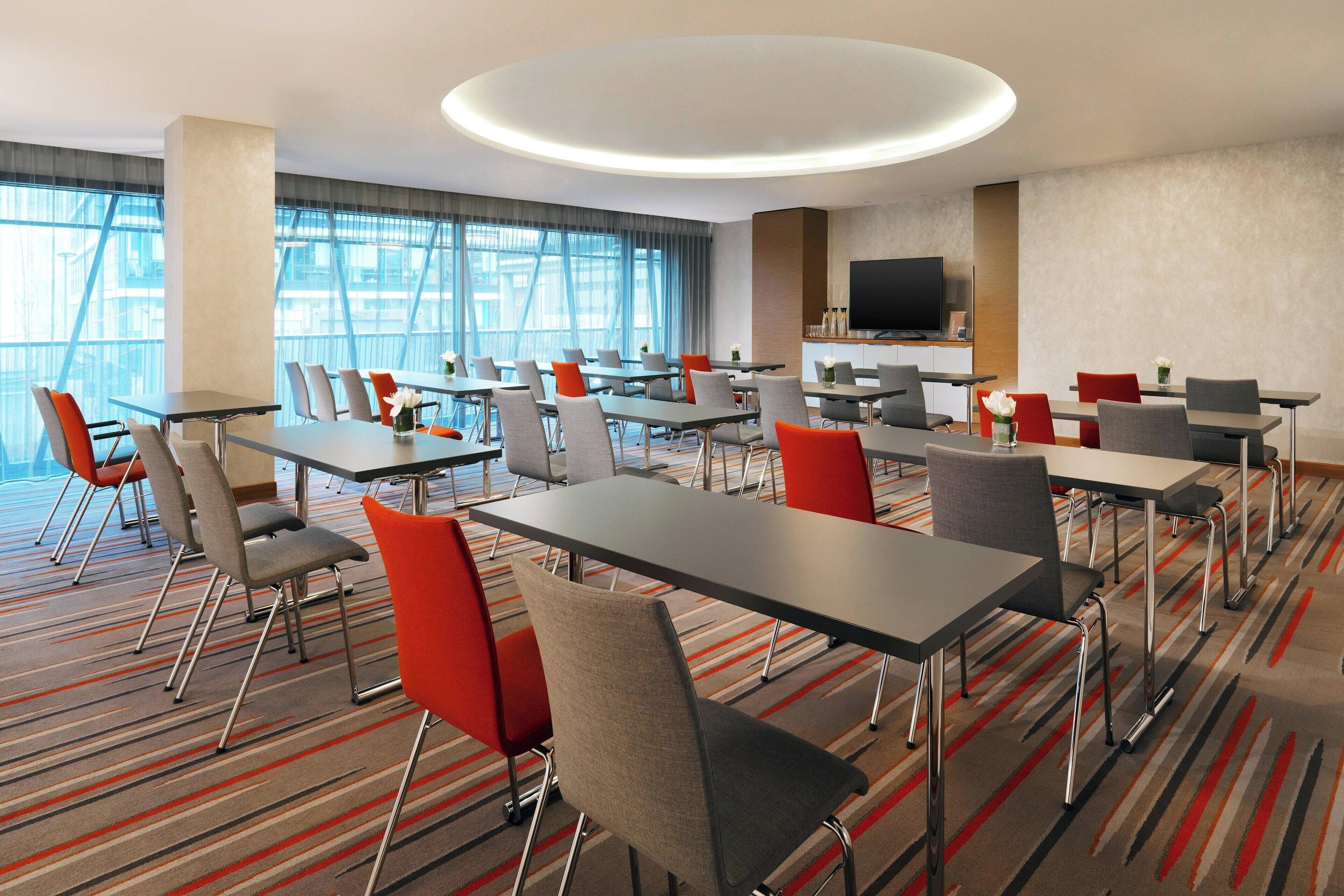 Een hotelconferentiezaal met meerdere tafels en stoelen, grote ramen, een flatscreen-televisie en een moderne plafondlamp.