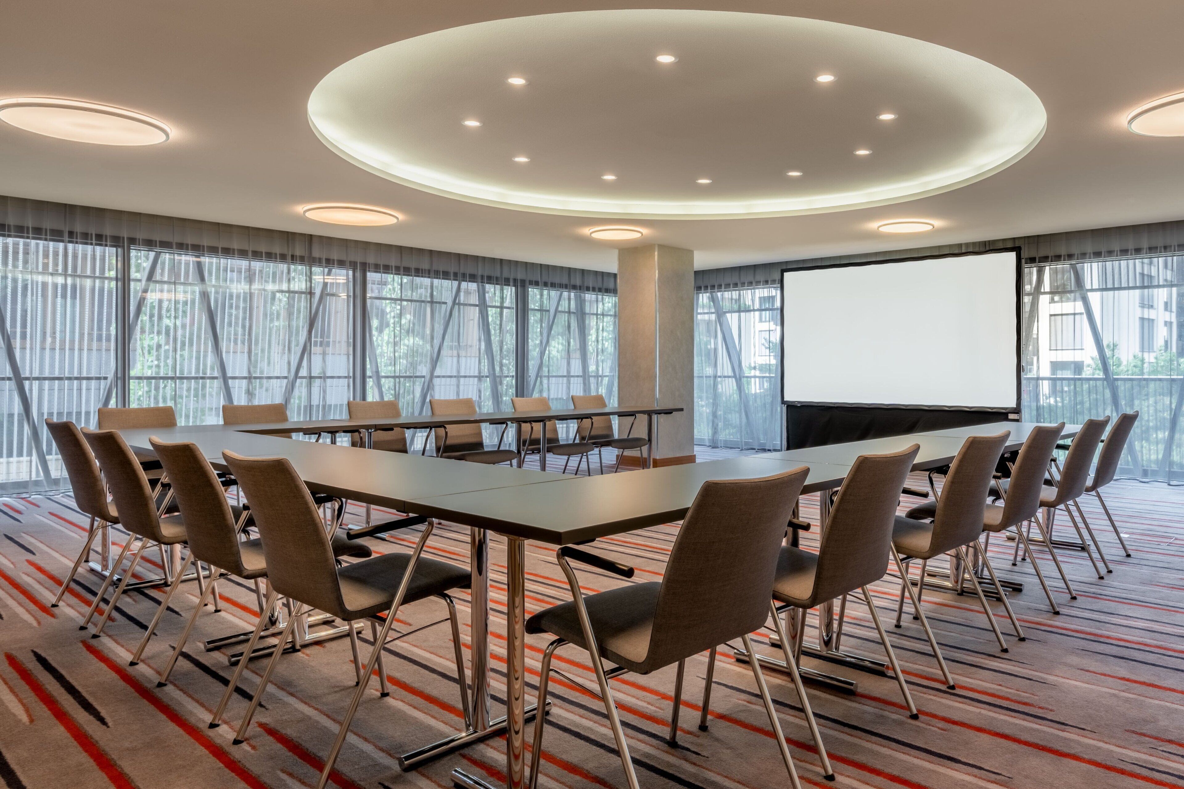 Un salón de conferencias con una gran mesa, sillas, una pantalla de proyector y un amplio ventanal con cortinas.