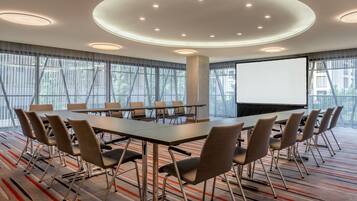 Un salón de conferencias con una gran mesa, sillas, una pantalla de proyector y un amplio ventanal con cortinas.