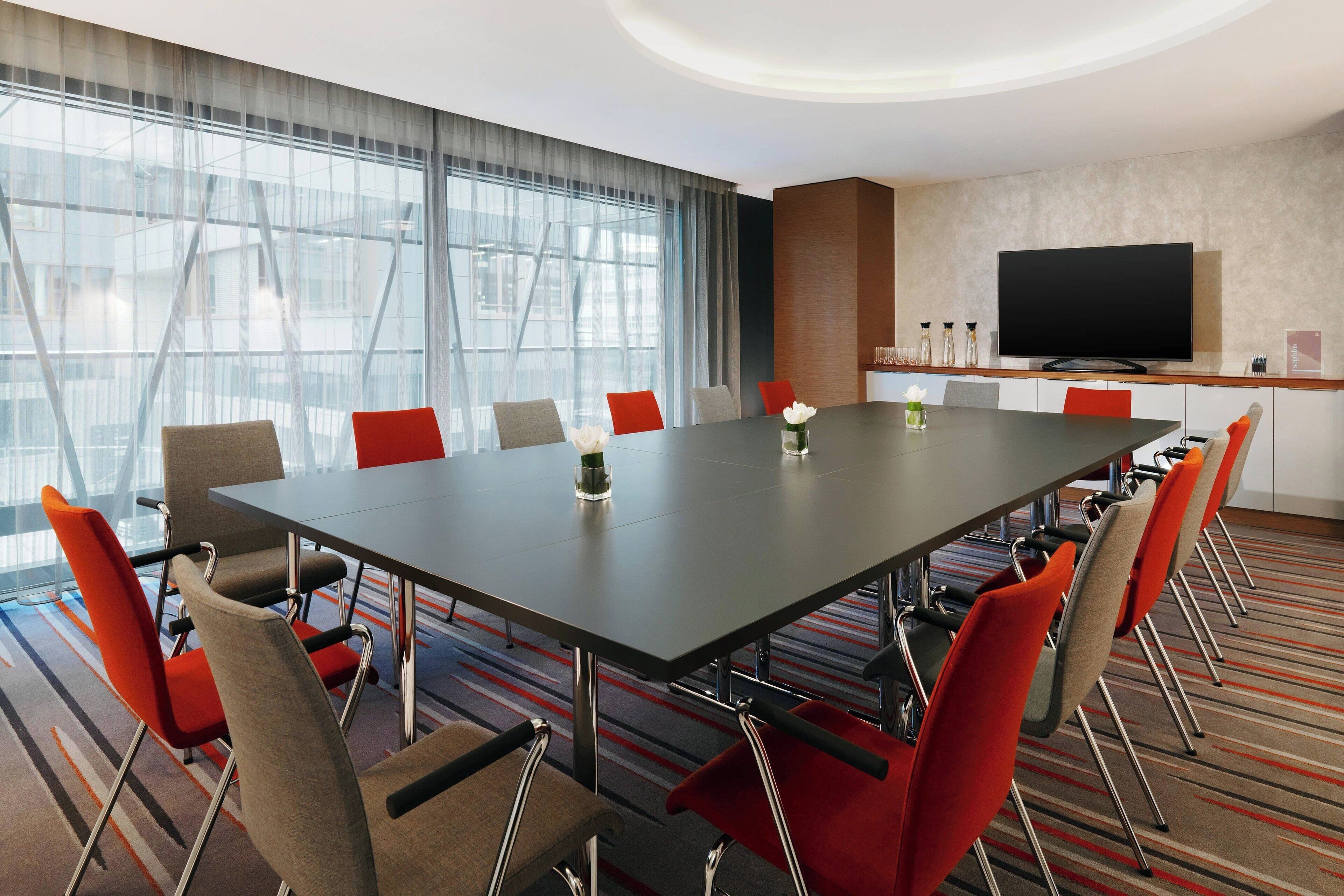 A conference room with a long table, red and grey chairs, a flat-screen TV, and a large window with curtains.
