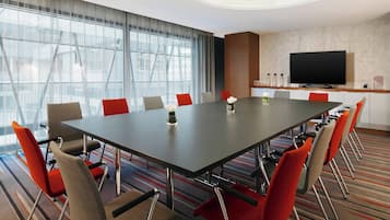 A conference room with a long table, red and grey chairs, a flat-screen TV, and a large window with curtains.