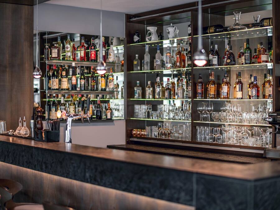 Uma área de bar com prateleiras cheias de garrafas de bebidas e copos, bancada de mármore e luminárias pendentes.