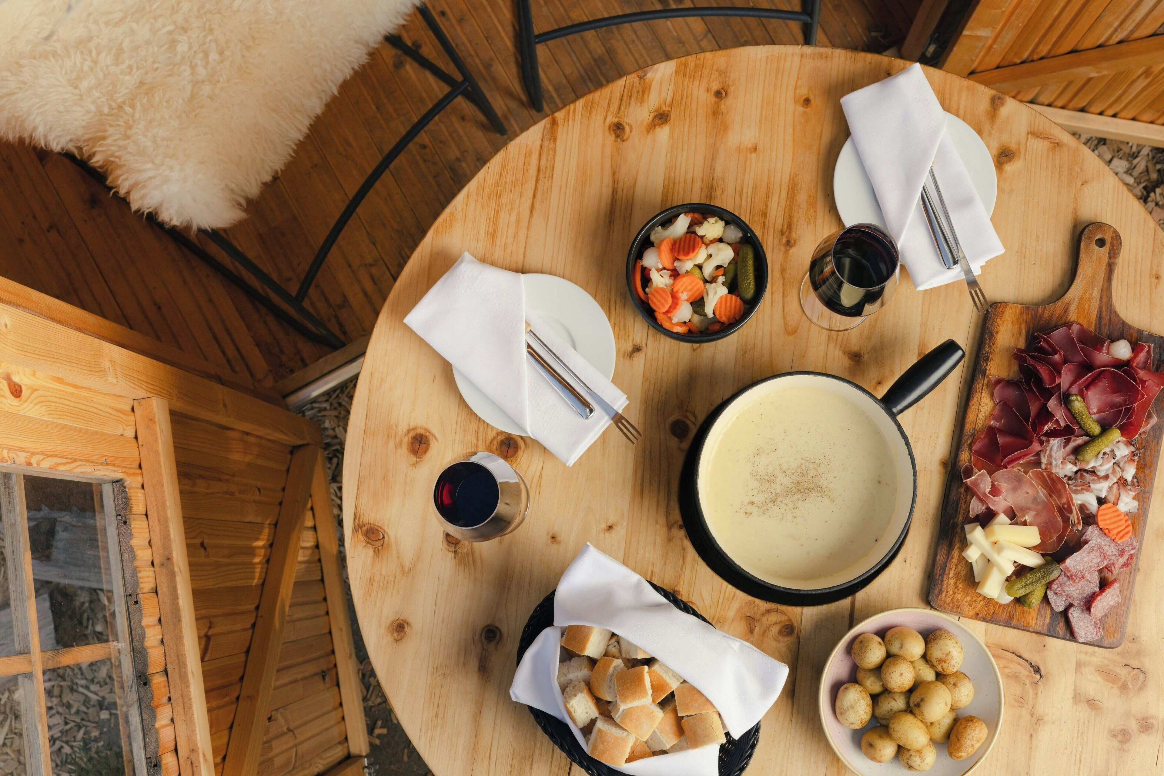 A round wooden table with a variety of food items, including a bowl of vegetables, a plate of meat, a basket of bread, and a bowl of potatoes.