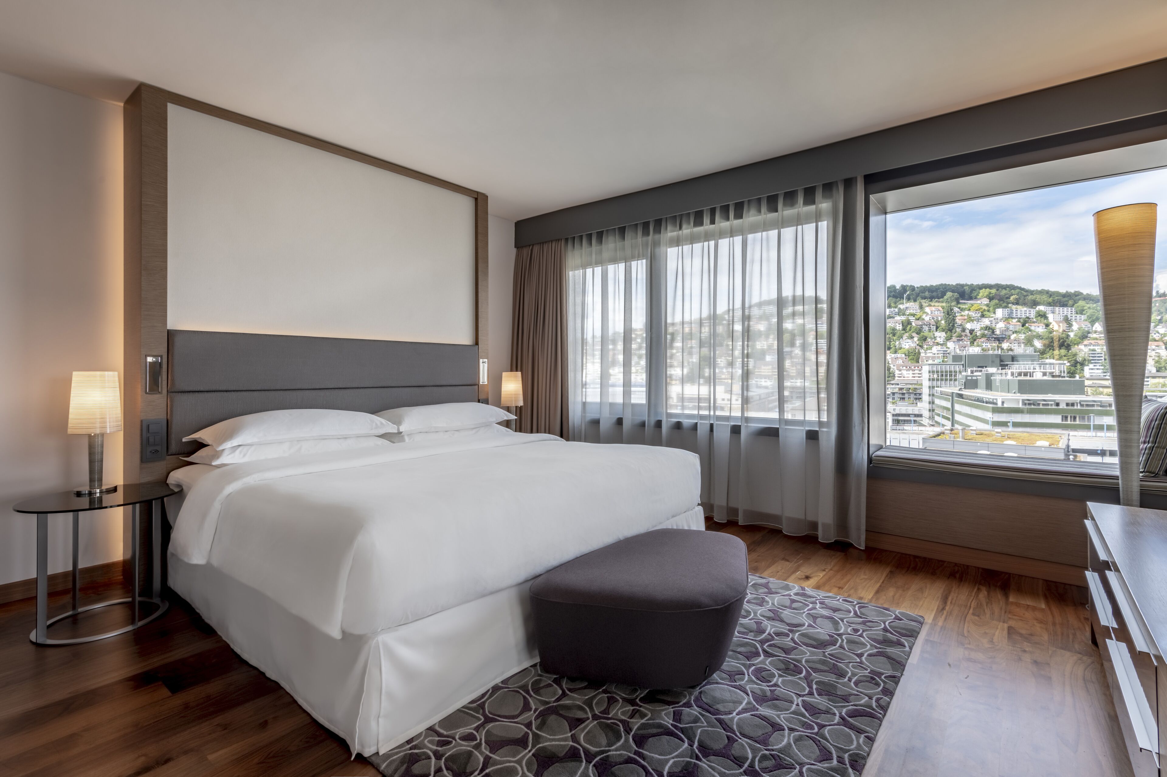 Ein modernes Hotelzimmer mit einem großen Bett, einem kleinen Tisch mit einer Lampe, einer Bank und Blick auf die Stadt durch große Fenster.