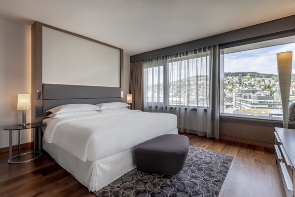 A modern hotel room with a large bed, a small table with a lamp, a bench, and a view of the city through large windows.