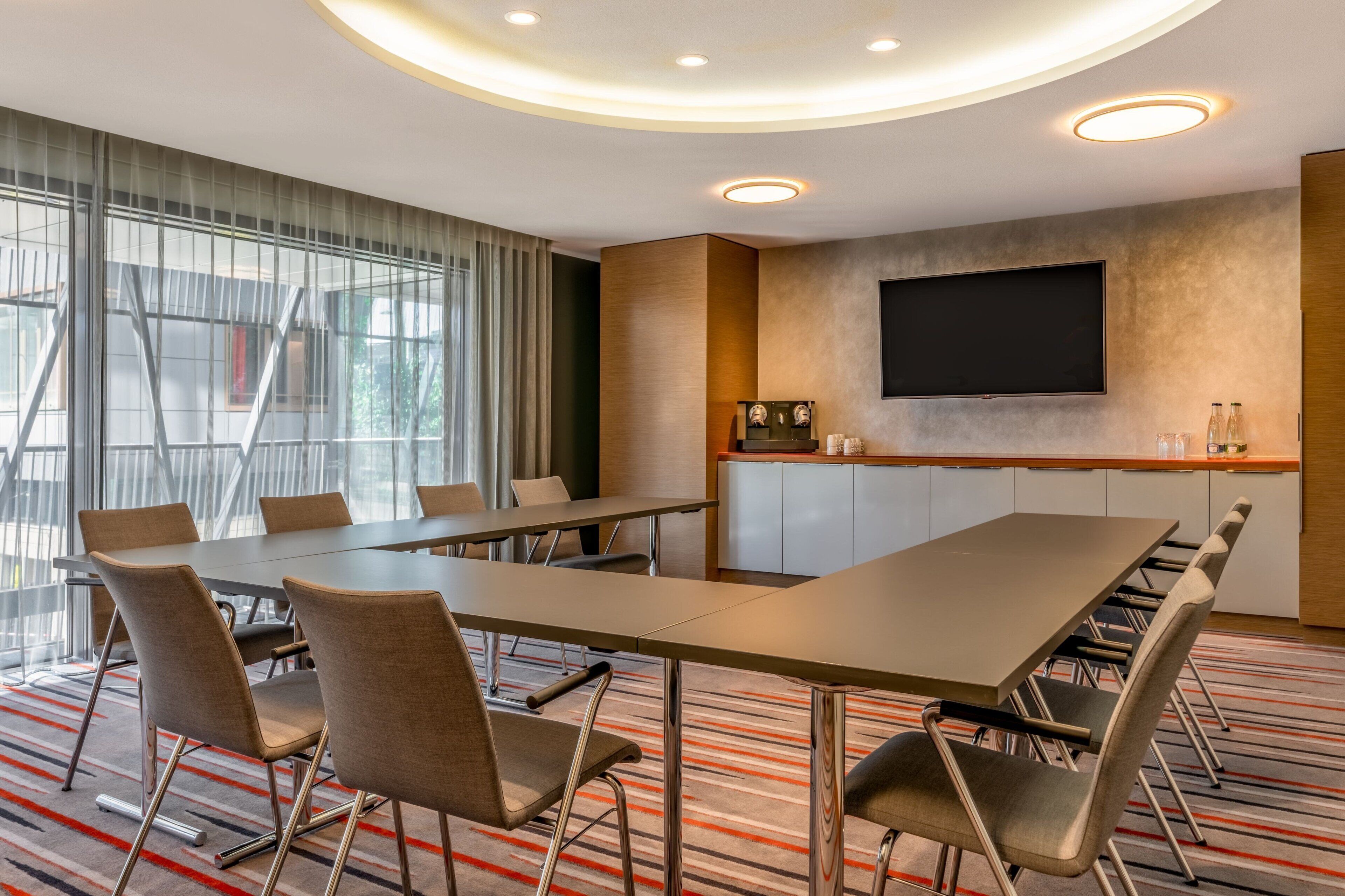 A conference room with a long table, chairs, a flat-screen TV, and a kitchen area with cabinets and a countertop.