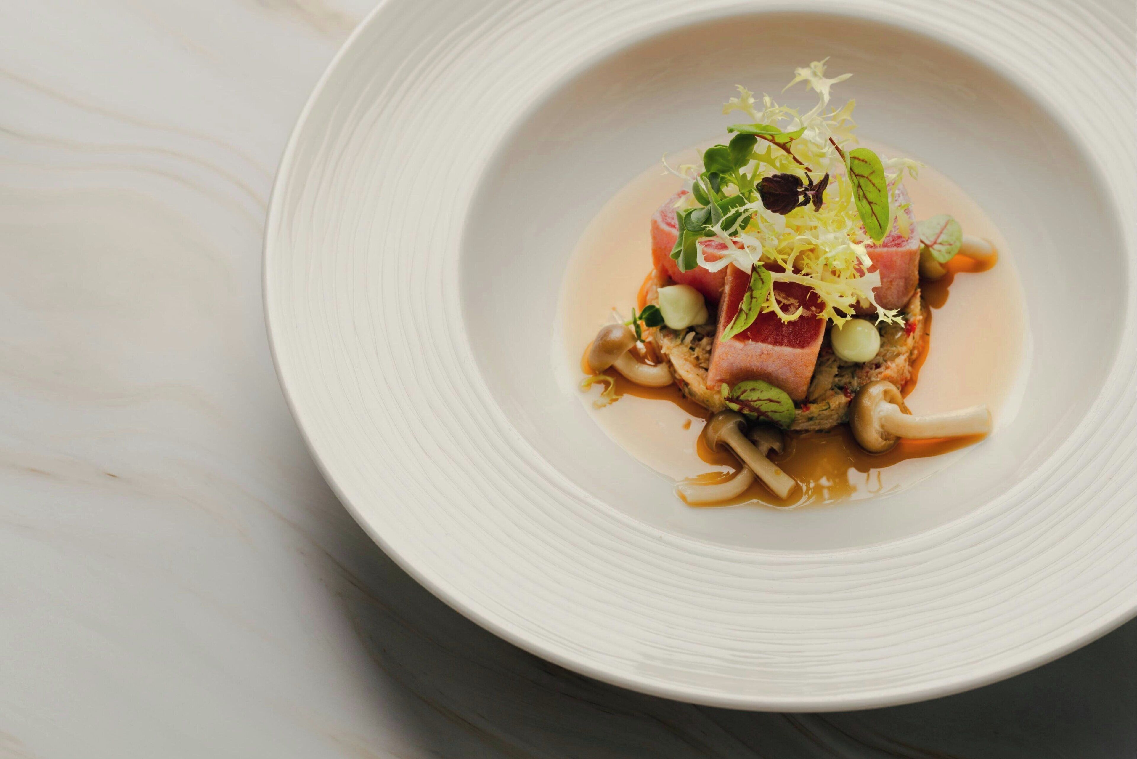 A white plate with a dish of food, including meat, mushrooms, and greens, on a marble surface.