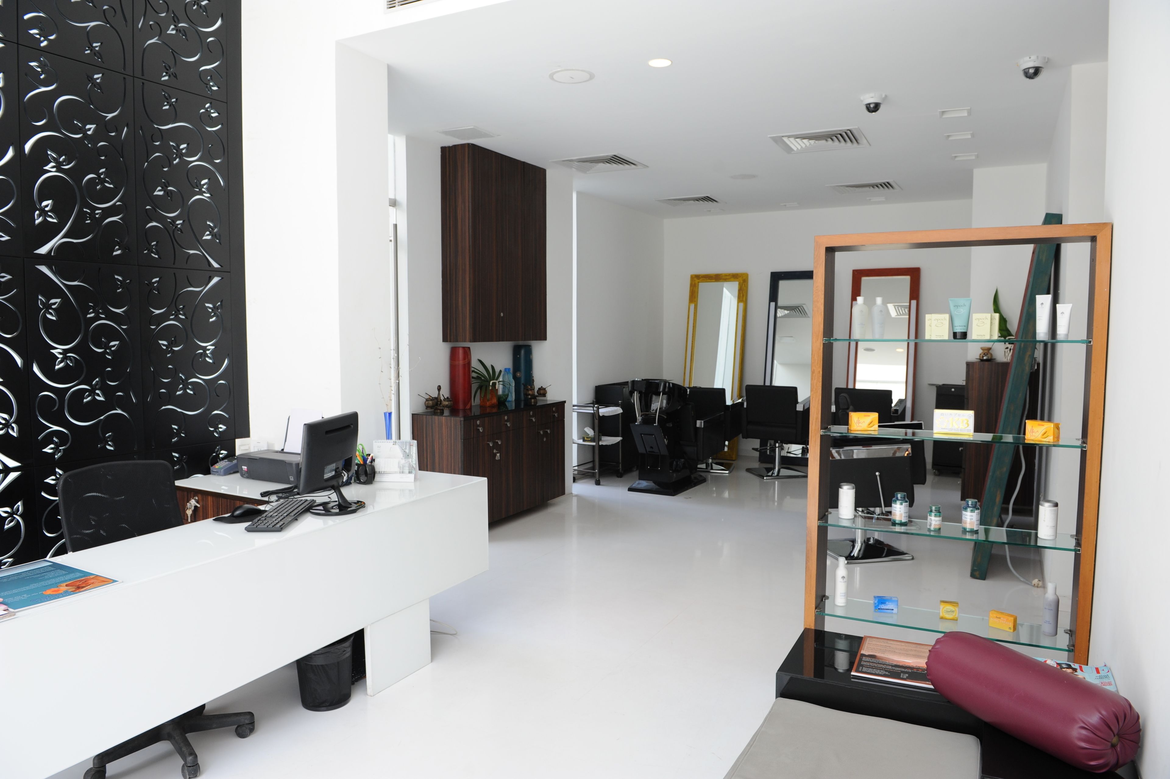 Oficina moderna con escritorio blanco, computadora y silla negra. Hay un espejo en la pared y una estantería con varios objetos.