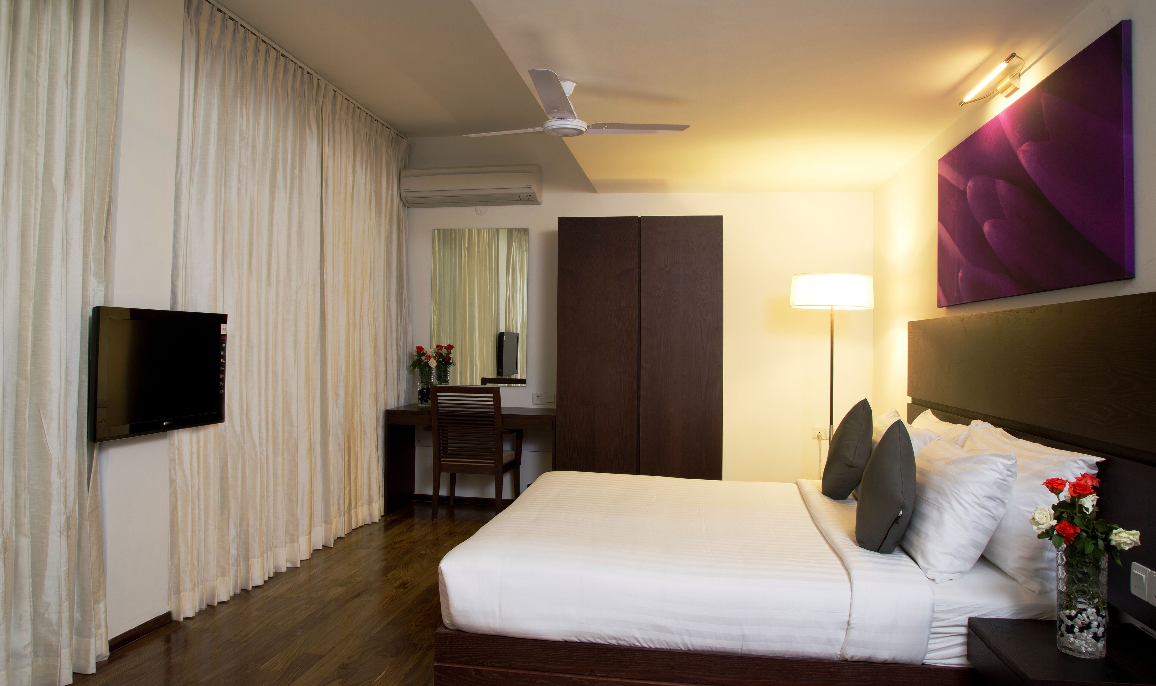 Une chambre d’hôtel comprenant un lit, un bureau, une chaise, une télévision, un ventilateur de plafond, une lampe et un vase avec des fleurs.
