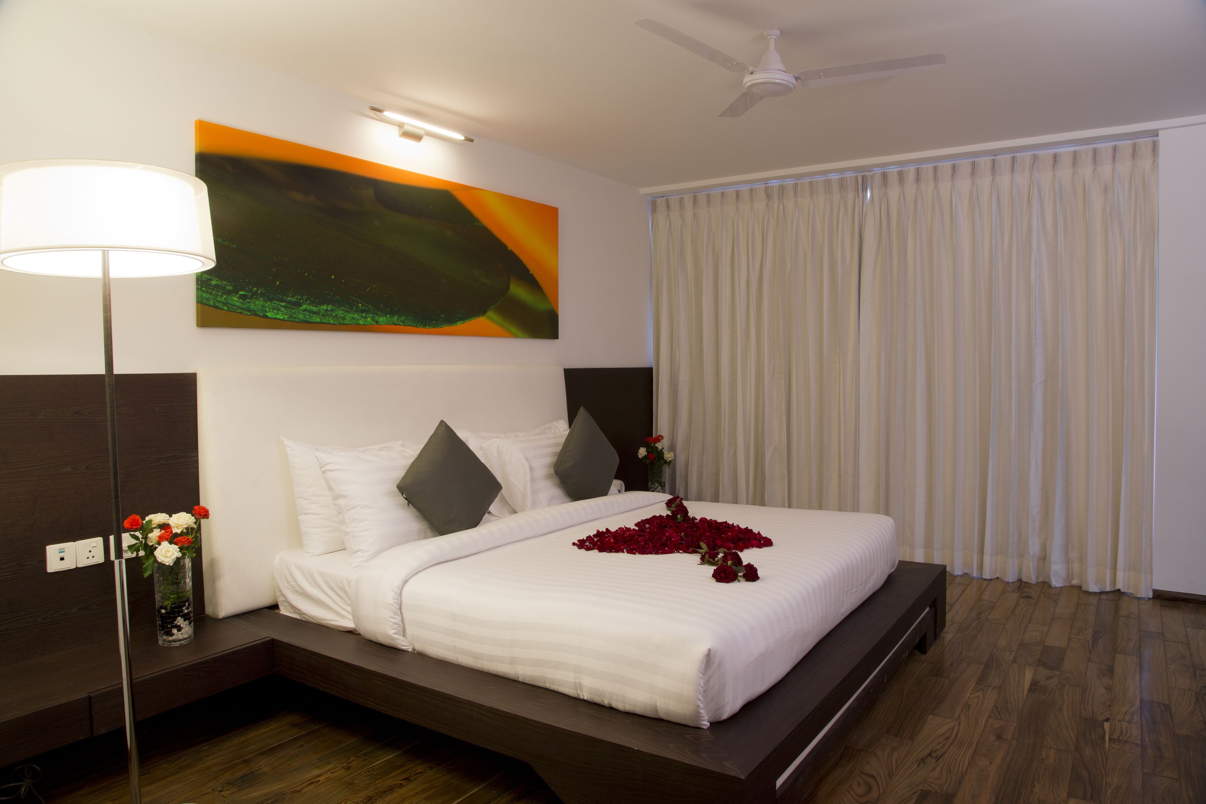 A modern hotel room with a large bed, a ceiling fan, a painting on the wall, and a window with curtains.