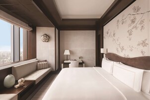 A modern hotel room with a large bed, a seating area, a desk, and a view of the city.