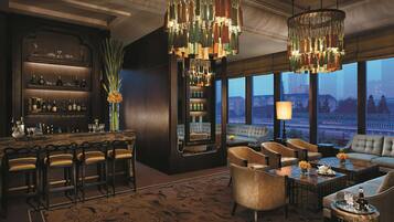 Eine Bar mit Bartresen, mehreren Barhocker und einer großen Chandelier. Eine Sitzecke mit Sofas und Sesseln, und ein großes Fenster mit Blick auf die Stadtlandschaft.