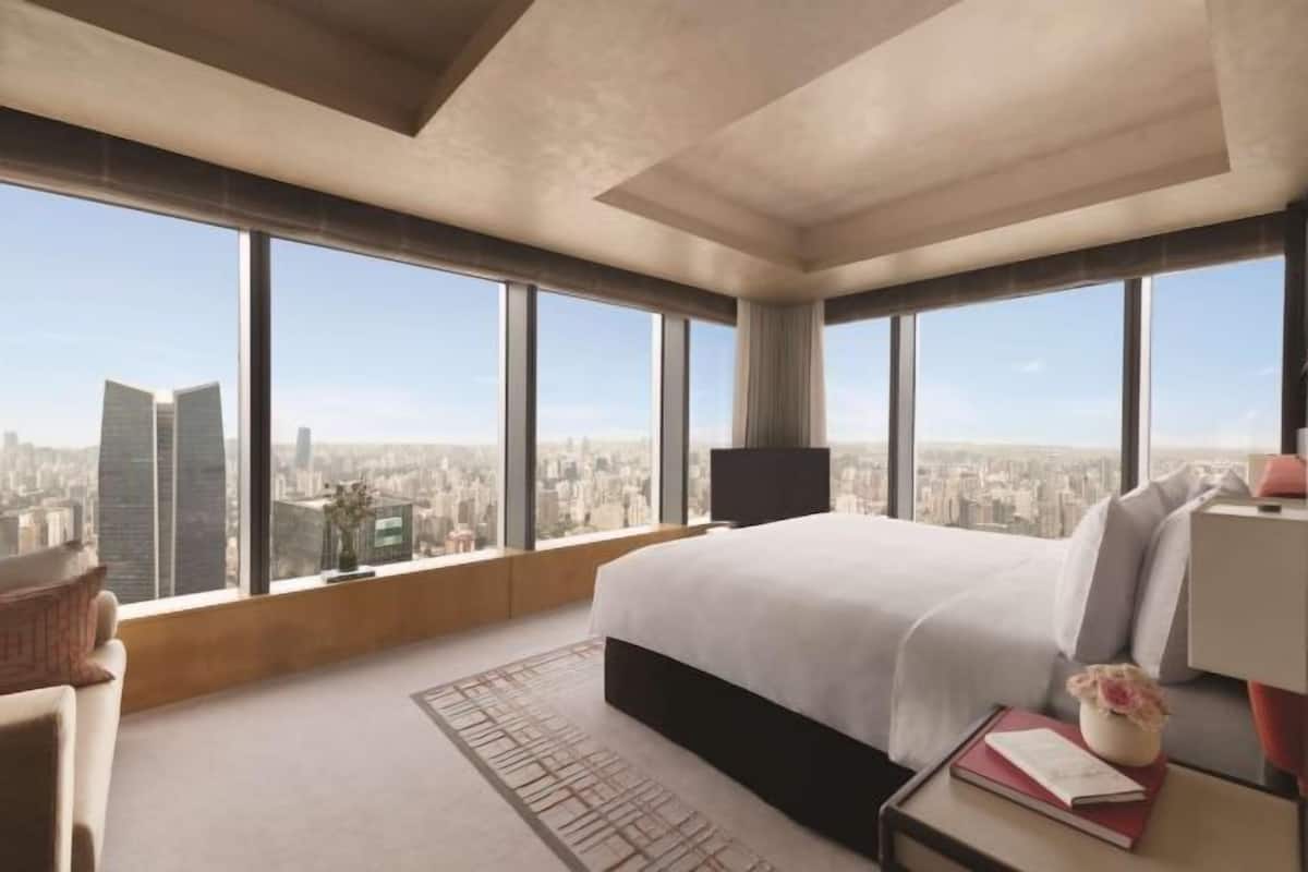 Habitación de hotel en un edificio alto con una cama grande, un televisor de pantalla plana, una mesita de noche y vistas de la ciudad.