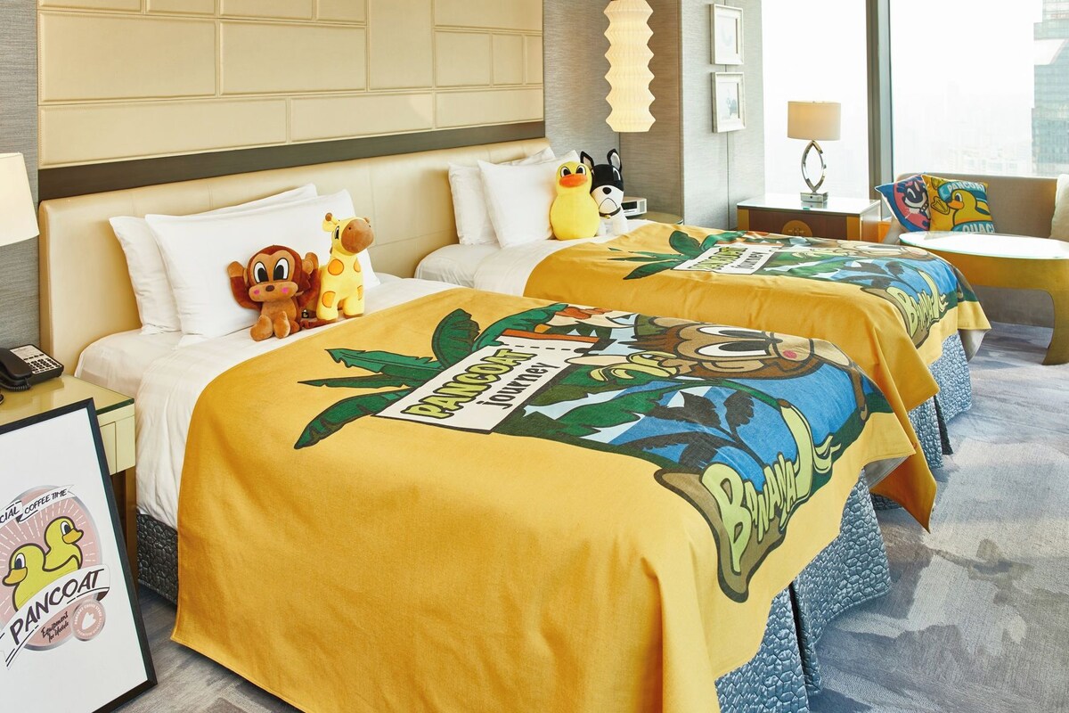 A hotel room with two beds, each with a yellow bedspread featuring a cartoon character and the word 'PANCOAT.' There are plush toys on the beds, a framed picture on the nightstand, and a lamp on the side table.