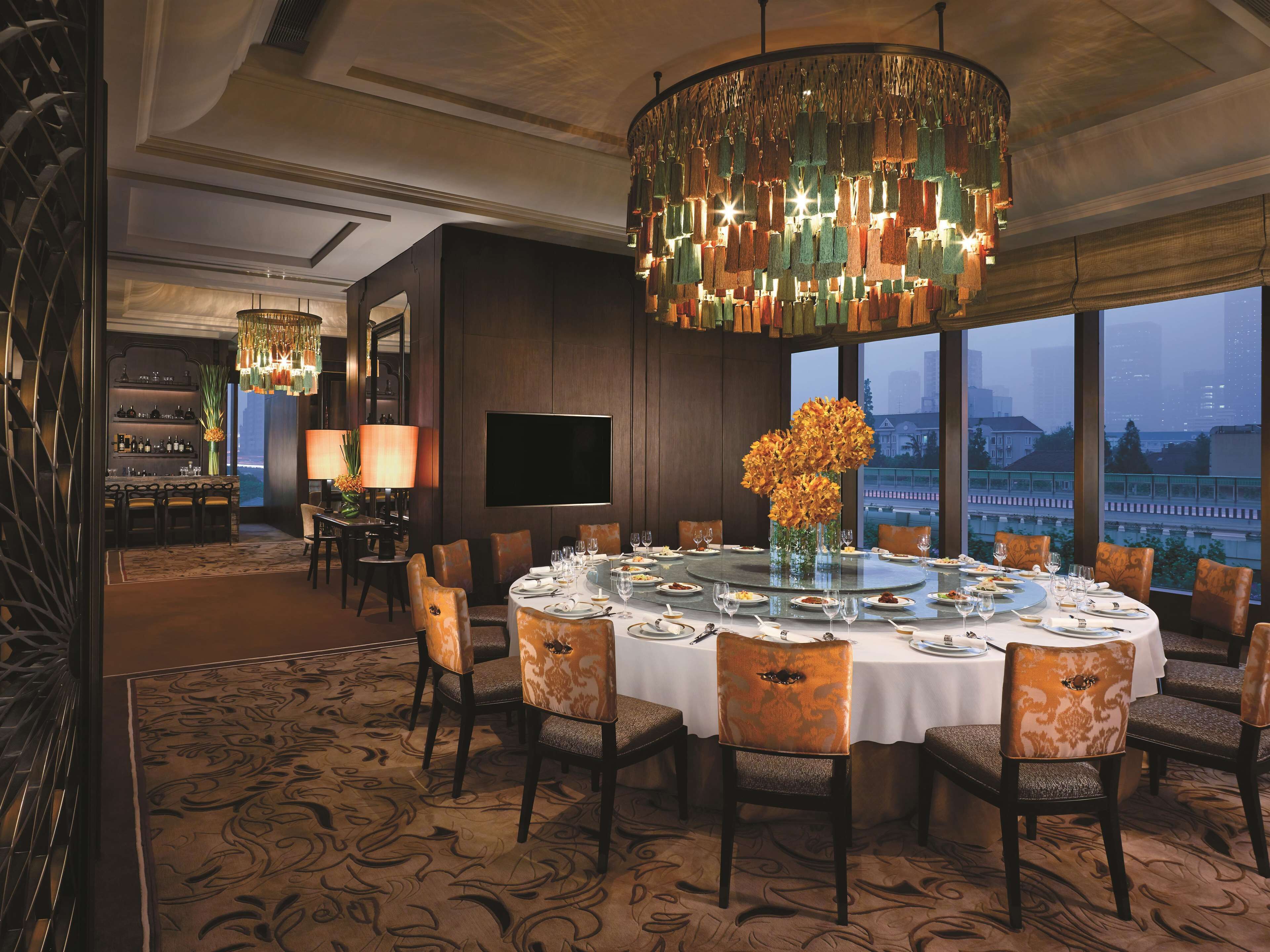 A dining area with a round table set for six, surrounded by chairs with patterned upholstery. A large, ornate chandelier hangs above the table, and a large window offers a view of the cityscape.
