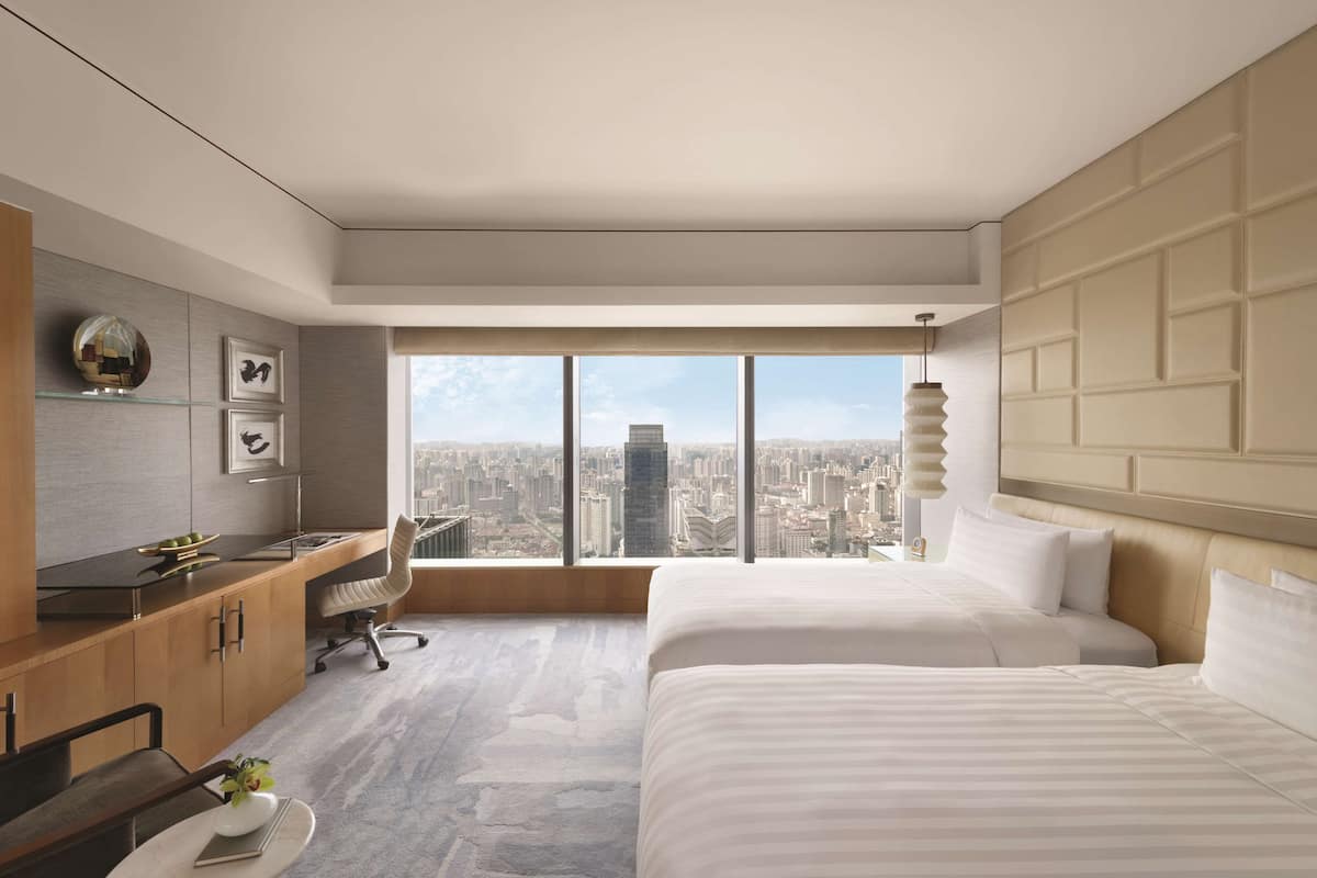 Una habitación de hotel moderna con un amplio ventanal, un escritorio, dos camas y vistas a la ciudad.