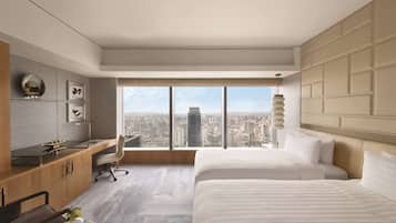 A modern hotel room with a large window, a desk, two beds, and a city view.