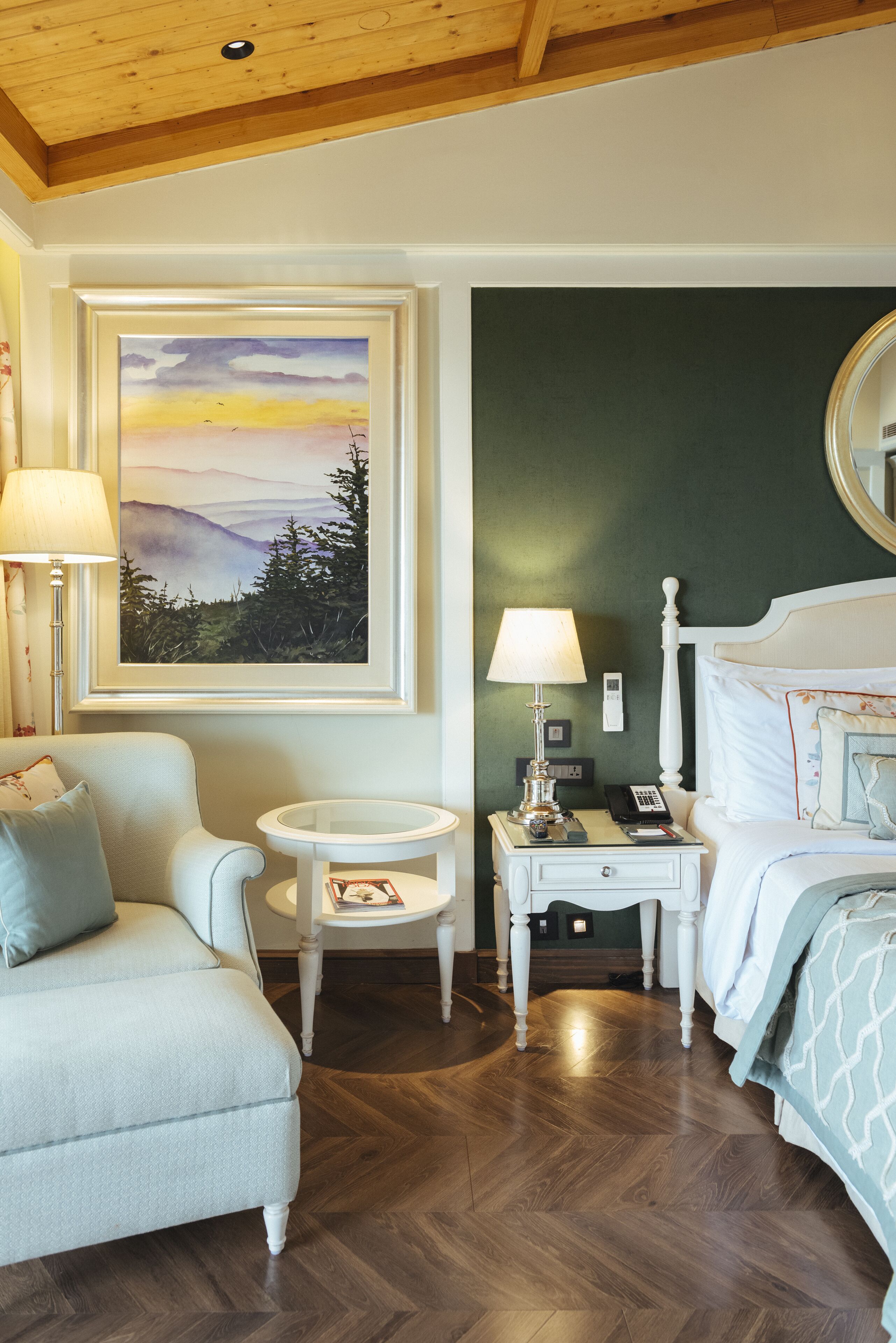 A hotel room with a wooden ceiling, a painting of a landscape, a bed with white bedding, a round table with a lamp, and a sofa.