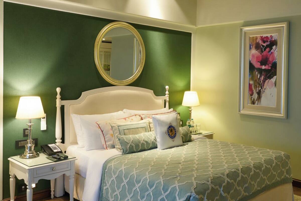 A neatly made bed with a green and white patterned bedspread, two bedside lamps, a round mirror, and a framed floral artwork on the wall.