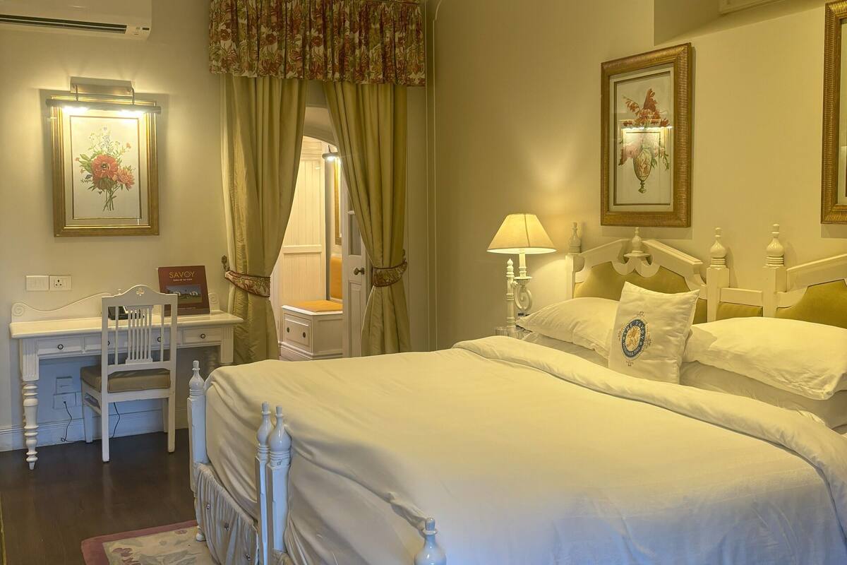 A double bed with white bedding, a desk with a chair, a bedside table with a lamp, and a framed picture on the wall.