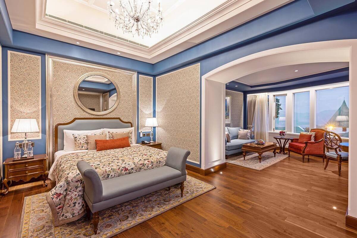 A spacious bedroom with a large bed, a sitting area with a sofa and armchairs, and a view of the cityscape through large windows.