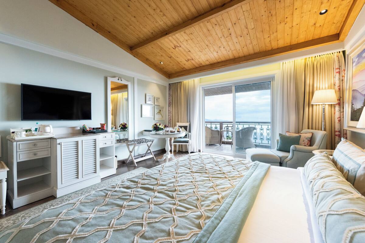 A hotel room with a large bed, a desk, a TV, and a balcony with a view.