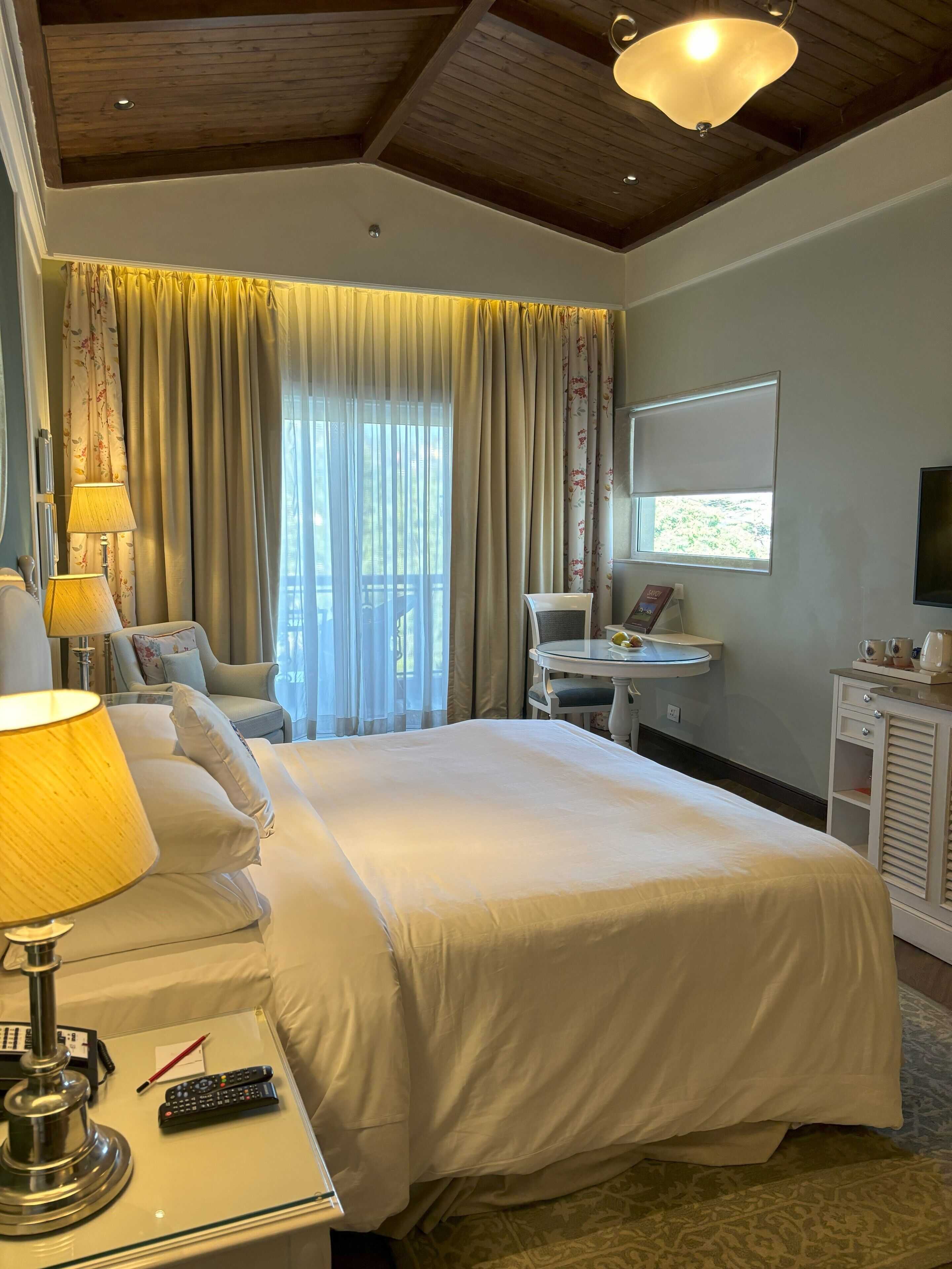 A hotel room with a large bed, a bedside table with a lamp, a desk with a chair, a television, and a window with curtains.