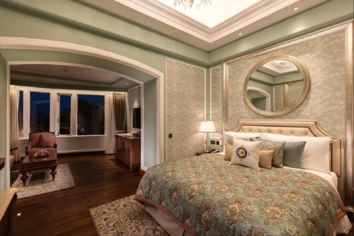 A spacious bedroom with a large bed, a sitting area with a chair and a table, a large window with curtains, and a mirror on the wall.