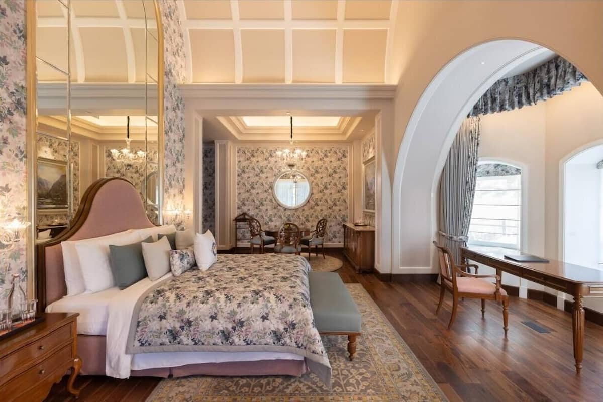 A bedroom with a large bed, a desk, and a chair. There is a chandelier and a window with curtains.