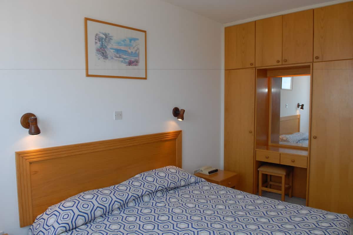 Ein Schlafzimmer mit Holzkopfstütze, einem Bett mit gemusterten Betttextilien, einem Nachttisch, einer Nachttischlampe, einem Holzschrank und einem gerahmten Bild an der Wand.