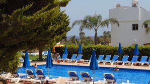 A pool area with lounge chairs and umbrellas, surrounded by greenery and a building in the background. - Maistros Hotel Apartments (Protaras)