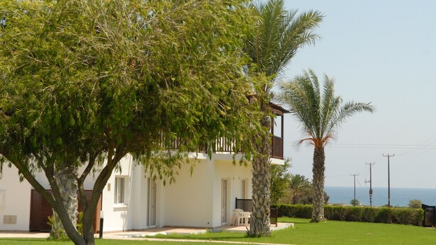 A white building with a balcony, surrounded by greenery and a clear view of the sea. - Maistros Hotel Apartments (Protaras)