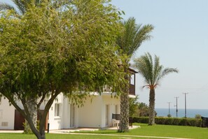 A white building with a balcony, surrounded by greenery and a clear view of the sea.