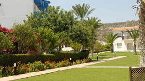 A well-maintained garden with a pathway, palm trees, and a building with blue accents. - Maistros Hotel Apartments (Protaras)