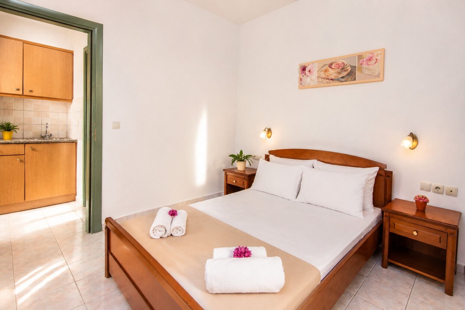 Una habitación de hotel con cama, mesitas de noche, una lámpara de pared, una pequeña planta en maceta y un cuadro.