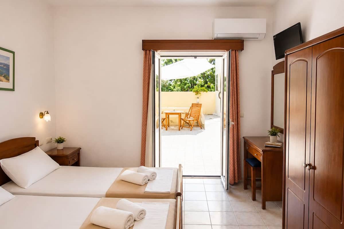 A hotel room with two beds, a desk, a wardrobe, and a view of an outdoor patio.