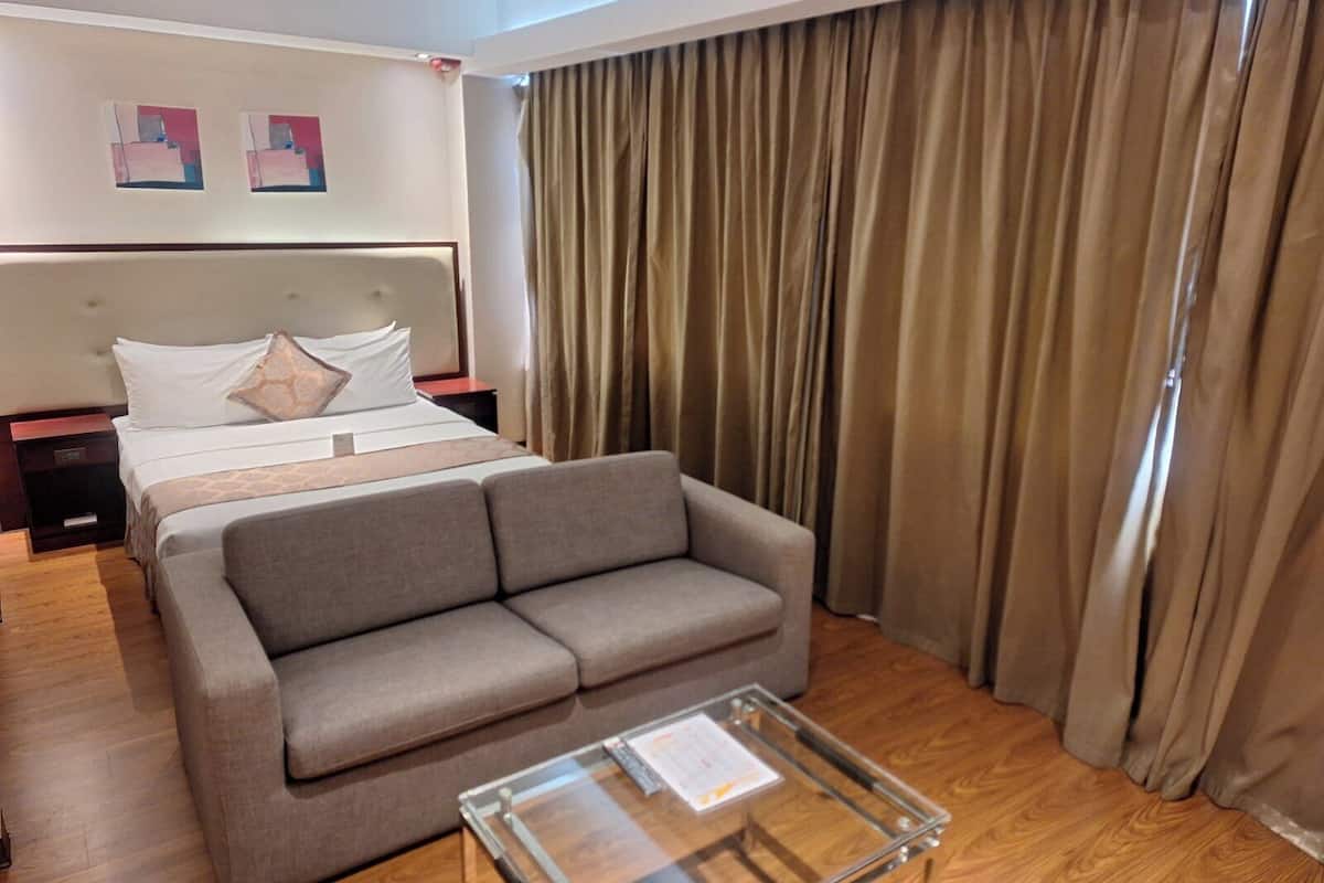 Ein Hotelzimmer mit einem Bett, einem grauen Sofa, einem Glastisch, Holzboden und beigen Vorhängen.
