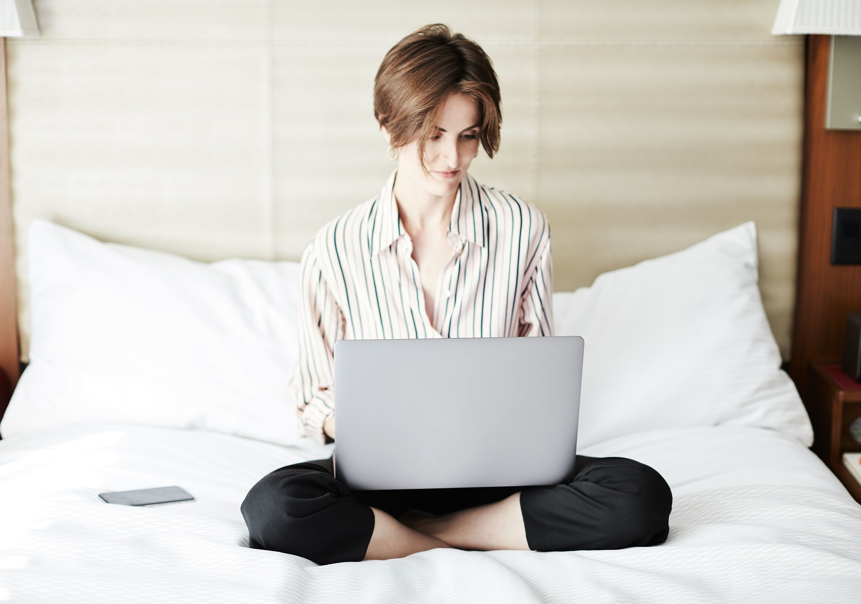 A person sitting on a bed with a laptop, using a smartphone, and a pillow is visible.