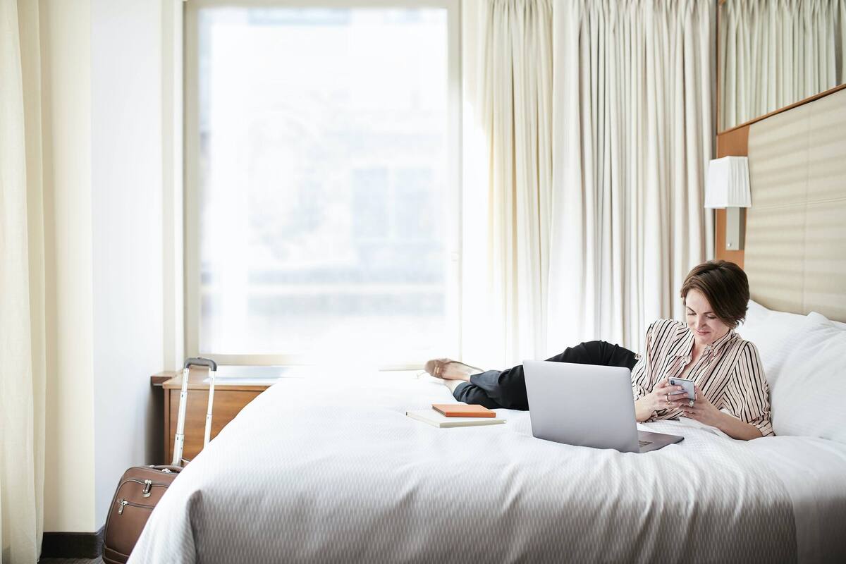Une chambre d’hôtel avec un lit, un ordinateur portable, une valise et une fenêtre avec des rideaux.